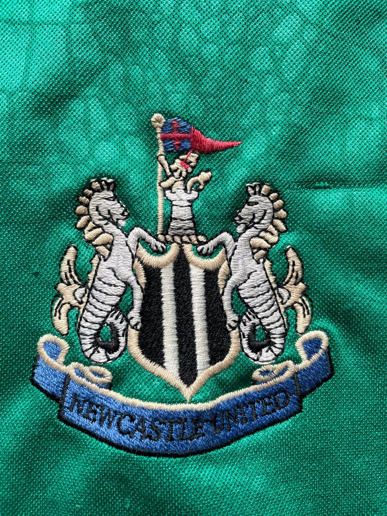 K&ouml;nnte beinhalten: Gesticktes Newcastle United Wappen auf gr&uuml;nem Stoff. Das Wappen zeigt zwei Seepferde, die einen Schild mit schwarz-wei&szlig;en Streifen flankieren. Ein Banner darunter lautet "NEWCASTLE UNITED". Oben befindet sich eine kleine Flagge.