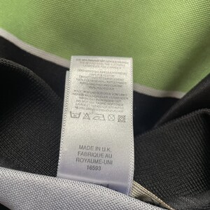 Pu&ograve; includere: Primo piano di un'etichetta di abbigliamento bianca con testo nero. L'etichetta include le istruzioni di lavaggio e il testo "MADE IN U.K. FABRIQUE AU ROYAUME-UNI 16593". Lo sfondo &egrave; verde chiaro.