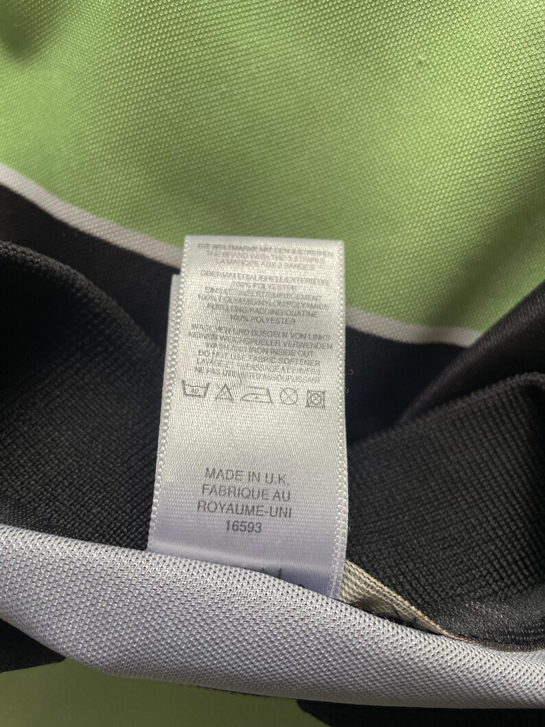 Pu&ograve; includere: Primo piano di un'etichetta di abbigliamento bianca con testo nero. L'etichetta include le istruzioni di lavaggio e il testo "MADE IN U.K. FABRIQUE AU ROYAUME-UNI 16593". Lo sfondo &egrave; verde chiaro.