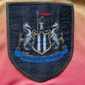 May include: Close-up of a Newcastle United patch. The patch is black with a shield shape and a brown border. The patch features the team's crest, including two seahorses, a flag, and the words "Newcastle United" in blue.