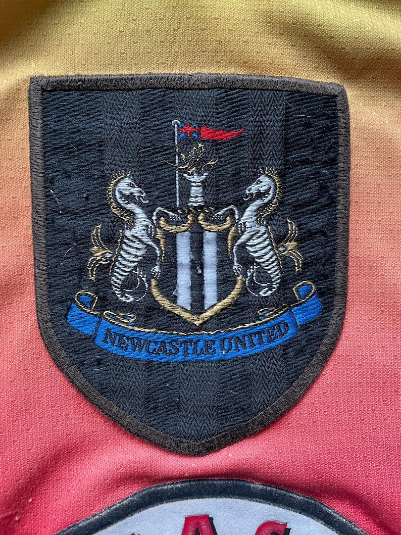 May include: Close-up of a Newcastle United patch. The patch is black with a shield shape and a brown border. The patch features the team's crest, including two seahorses, a flag, and the words "Newcastle United" in blue.