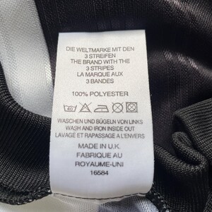 May include: A close-up of a garment label with text in German, English, and French. The label reads "THE BRAND WITH THE 3 STRIPES" and "100% POLYESTER". It also states the garment was made in the U.K. The garment has black and white stripes.