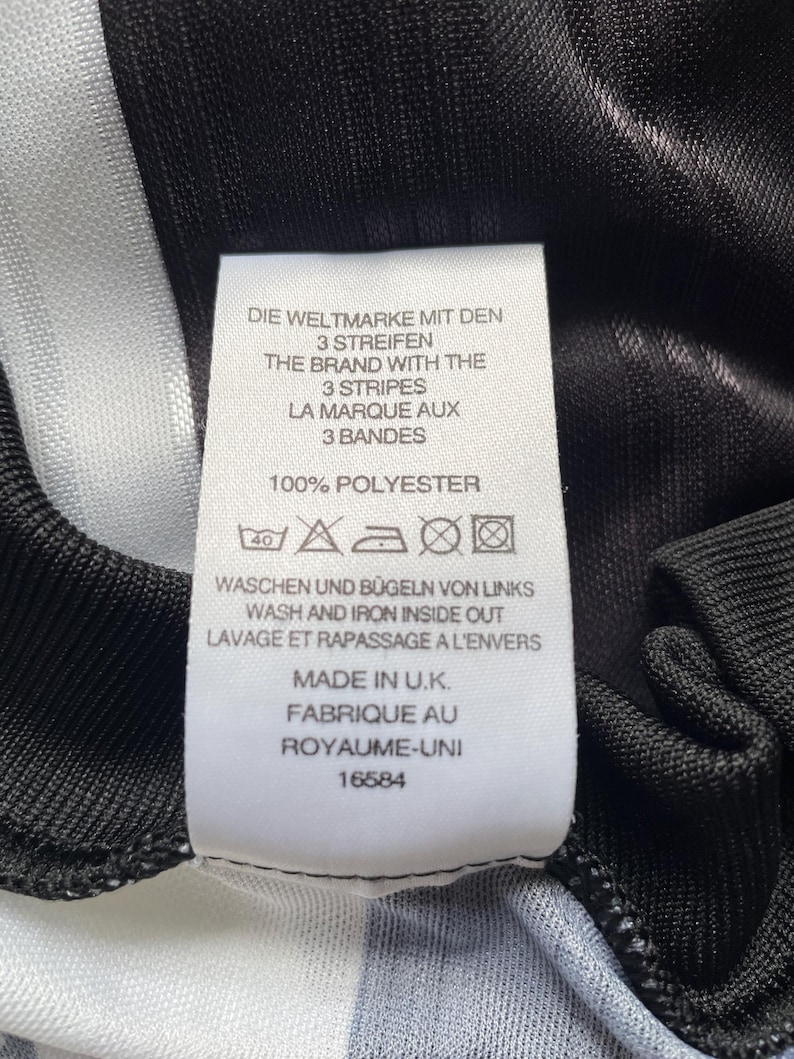May include: A close-up of a garment label with text in German, English, and French. The label reads "THE BRAND WITH THE 3 STRIPES" and "100% POLYESTER". It also states the garment was made in the U.K. The garment has black and white stripes.