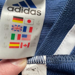 Op de afbeelding: Close-up van een marineblauw Adidas-label. Het label toont het Adidas-logo en vlaggen van verschillende landen, waaronder Duitsland, Japan, Groot-Brittanni&euml;, Frankrijk, Itali&euml;, de VS, Spanje en Canada. Maat- en stijlinformatie is ook zichtbaar.