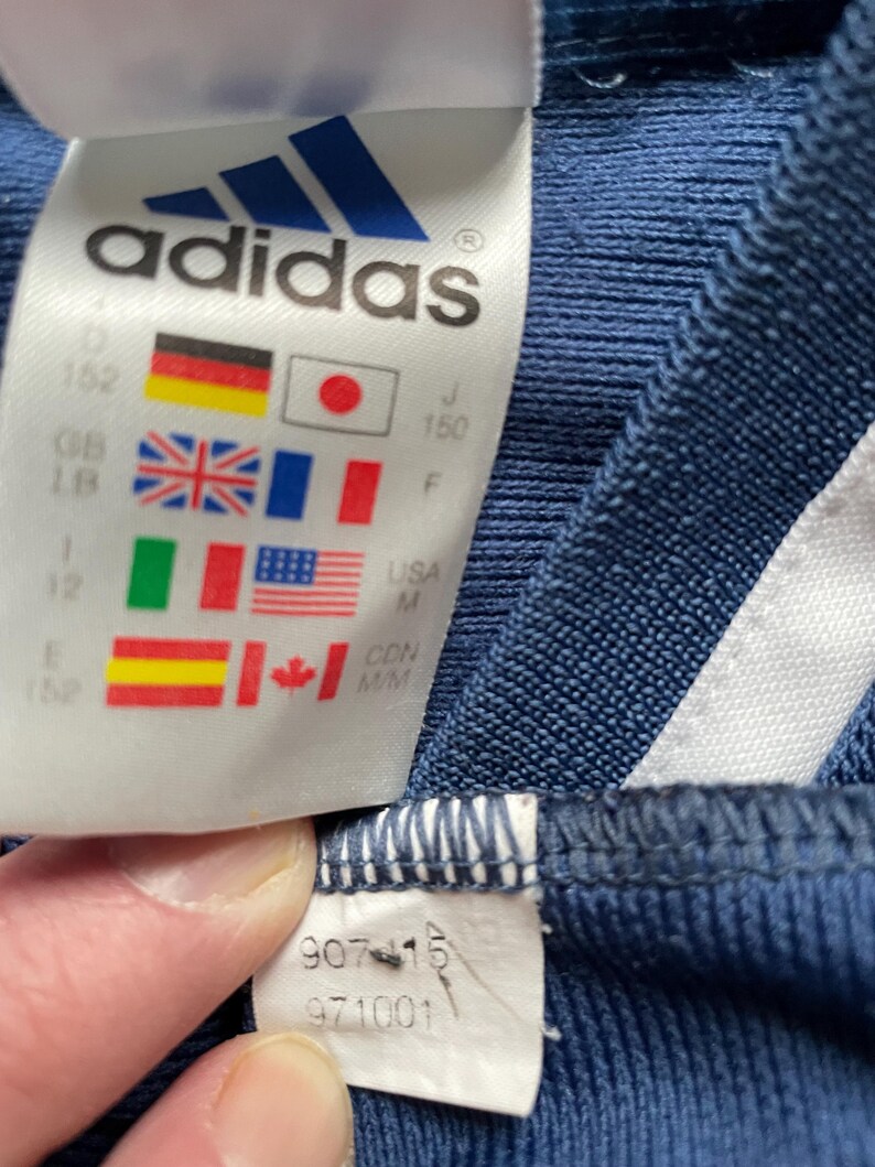 Op de afbeelding: Close-up van een marineblauw Adidas-label. Het label toont het Adidas-logo en vlaggen van verschillende landen, waaronder Duitsland, Japan, Groot-Brittanni&euml;, Frankrijk, Itali&euml;, de VS, Spanje en Canada. Maat- en stijlinformatie is ook zichtbaar.