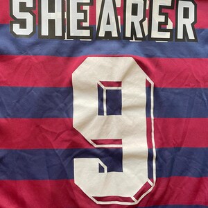 May include: Close-up of a sports jersey with the name "SHEARER" in white block letters across the top. Below, a large white number "9" is centered. The jersey has alternating red and blue horizontal stripes.