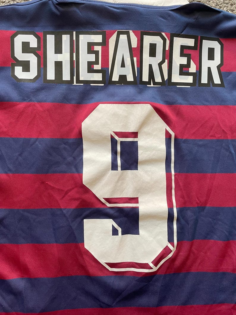May include: Close-up of a sports jersey with the name "SHEARER" in white block letters across the top. Below, a large white number "9" is centered. The jersey has alternating red and blue horizontal stripes.