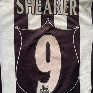 May include: A black and white striped soccer jersey with the name "SHEARER" across the upper back. The number "9" is prominently displayed on the front, within a shield-shaped emblem. The Premier League logo is visible.