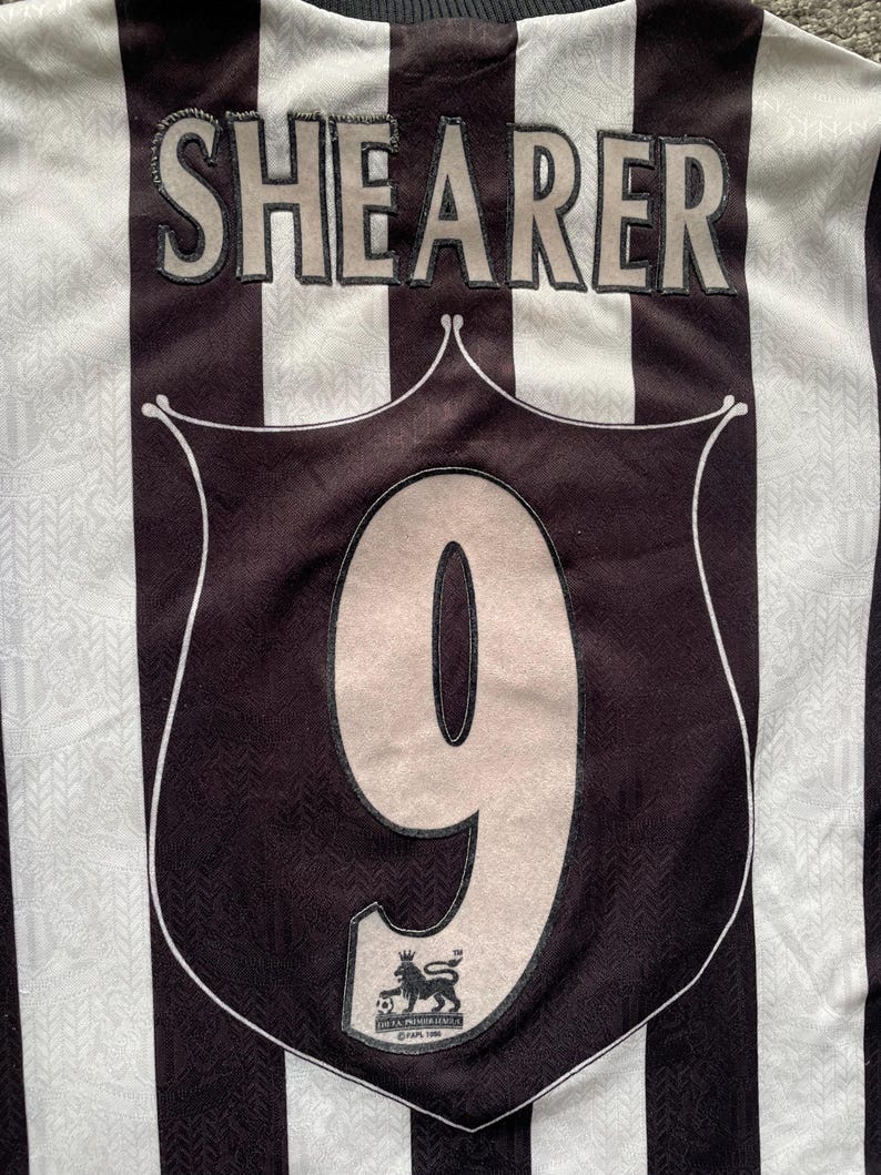 May include: A black and white striped soccer jersey with the name "SHEARER" across the upper back. The number "9" is prominently displayed on the front, within a shield-shaped emblem. The Premier League logo is visible.