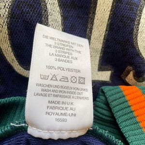 May include: Close-up of a navy blue garment with a white tag. The tag features text in multiple languages, including "THE BRAND WITH THE 3 STRIPES" and washing instructions. The garment has green and orange striped detailing.