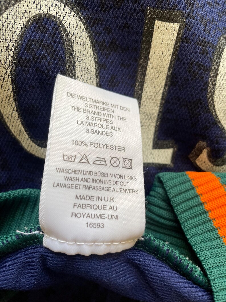 May include: Close-up of a navy blue garment with a white tag. The tag features text in multiple languages, including "THE BRAND WITH THE 3 STRIPES" and washing instructions. The garment has green and orange striped detailing.