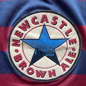 May include: A close-up of a striped shirt in blue and maroon. The shirt has a circular patch with the words "Newcastle Brown Ale" and a blue star with a city silhouette.