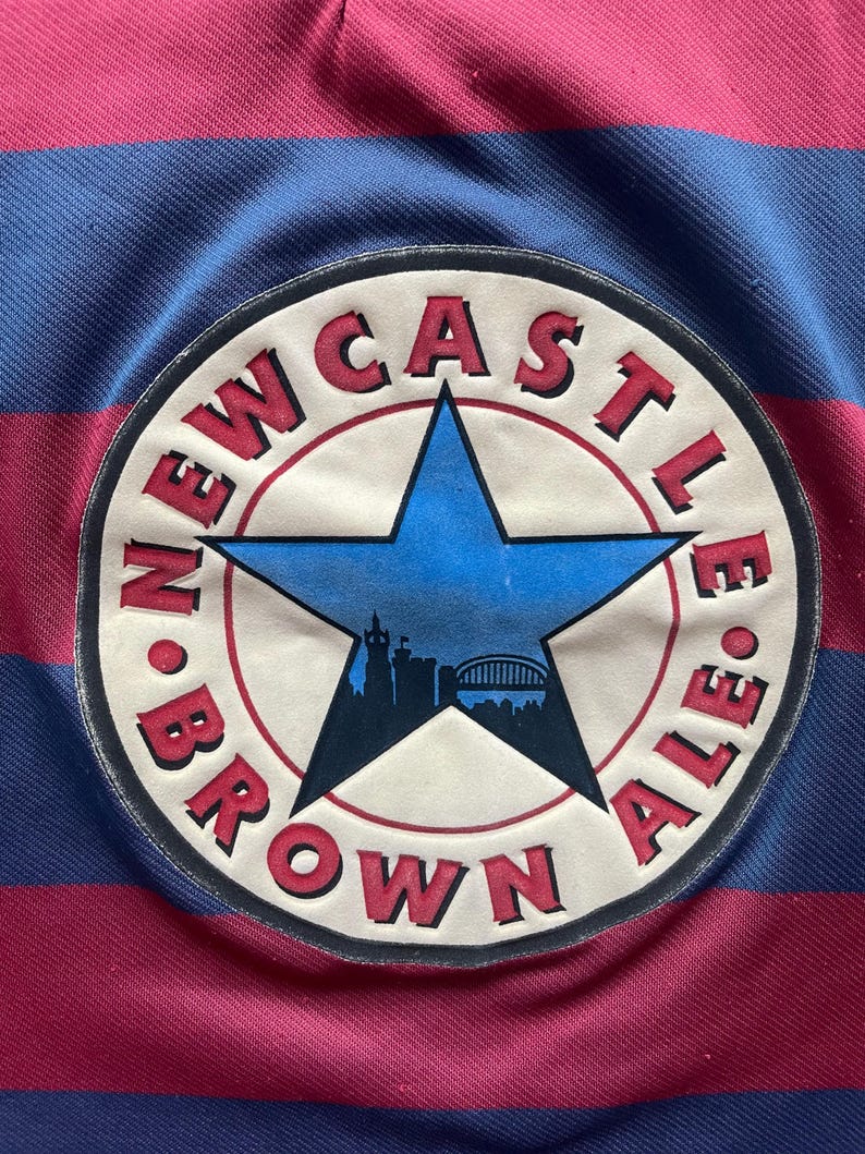 May include: A close-up of a striped shirt in blue and maroon. The shirt has a circular patch with the words "Newcastle Brown Ale" and a blue star with a city silhouette.