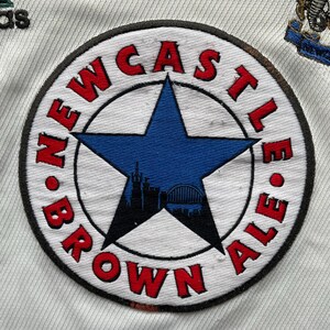 Puede incluir: Camiseta de f&uacute;tbol blanca con un parche circular. El parche dice "NEWCASTLE BROWN ALE" en rojo alrededor de una estrella azul, con una silueta de la ciudad. Logotipo de Adidas visible.