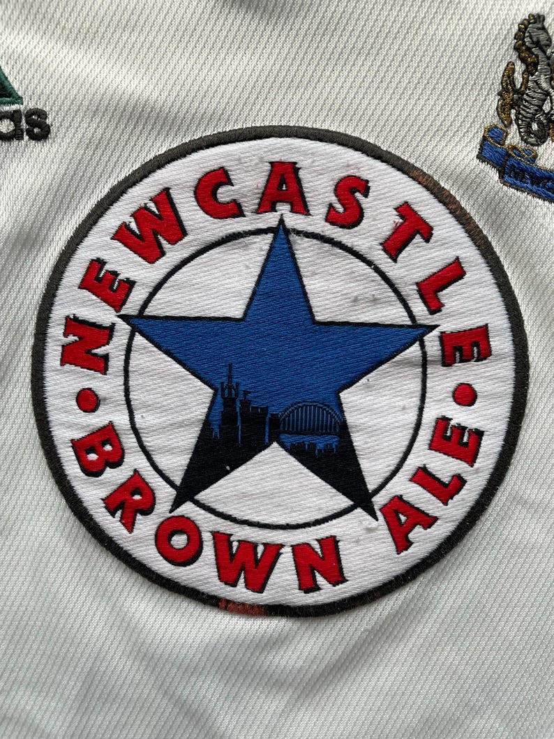 Puede incluir: Camiseta de f&uacute;tbol blanca con un parche circular. El parche dice "NEWCASTLE BROWN ALE" en rojo alrededor de una estrella azul, con una silueta de la ciudad. Logotipo de Adidas visible.