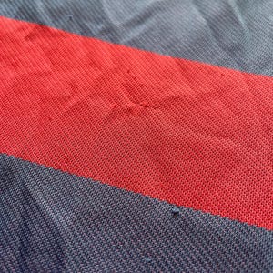 May include: Close-up of a red, navy blue, and light blue striped fabric. The fabric appears to be a knit material, with visible texture and slight imperfections. The stripes are wide and run horizontally across the frame.