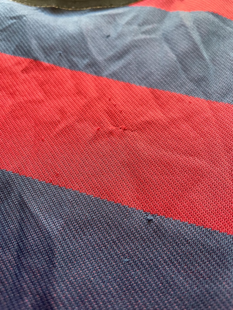 May include: Close-up of a red, navy blue, and light blue striped fabric. The fabric appears to be a knit material, with visible texture and slight imperfections. The stripes are wide and run horizontally across the frame.