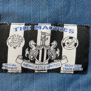 May include: Black and white patch with the text "The Magpies" and a crest featuring a black and white bird with a shield and two seahorses. The text "Official Garment" and "Newcastle United" are also visible.