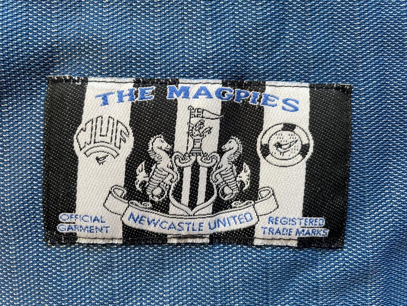 May include: Black and white patch with the text "The Magpies" and a crest featuring a black and white bird with a shield and two seahorses. The text "Official Garment" and "Newcastle United" are also visible.