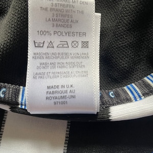 May include: A white care label with black text detailing the fabric composition and care instructions for a garment. The label reads "100% Polyester" and includes washing and ironing instructions in German, English, and French. The label also states "Made in U.K. Fabrique au Royaume-Uni 971001".