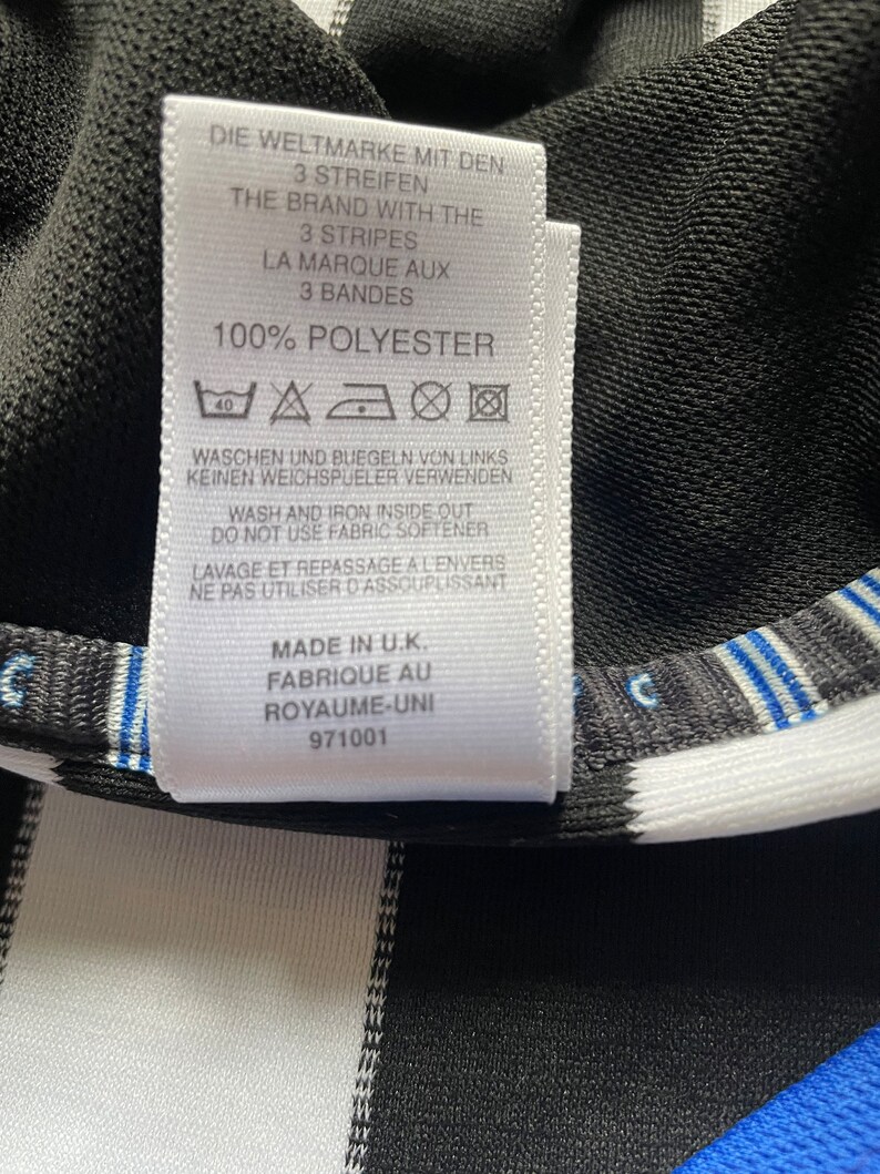 May include: A white care label with black text detailing the fabric composition and care instructions for a garment. The label reads "100% Polyester" and includes washing and ironing instructions in German, English, and French. The label also states "Made in U.K. Fabrique au Royaume-Uni 971001".
