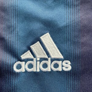 May include: Close-up of a blue and purple fabric with the Adidas logo embroidered in white. The fabric has a subtle striped pattern and a textured appearance, with the Adidas logo prominently displayed.