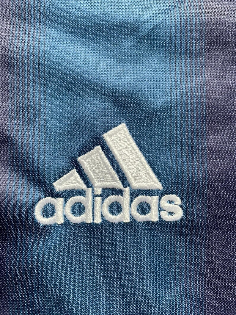 May include: Close-up of a blue and purple fabric with the Adidas logo embroidered in white. The fabric has a subtle striped pattern and a textured appearance, with the Adidas logo prominently displayed.