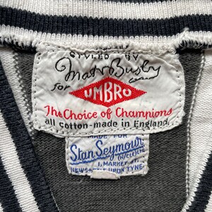 Puede incluir: Una etiqueta blanca con texto negro que dice "Styled by MatrBusby for Umbro The Choice of Champions all cotton - made in England" y un logotipo rojo que dice "Umbro". Debajo de la etiqueta hay una etiqueta blanca m&aacute;s peque&ntilde;a con texto azul que dice "Made for Stan Seymour (N/CLE)LYA 1. MARKET ST. NEWCASTLE UPON TYNE".