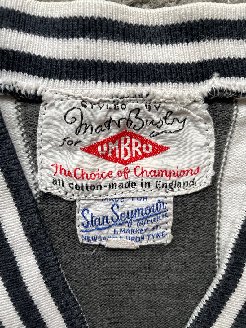 Puede incluir: Una etiqueta blanca con texto negro que dice "Styled by MatrBusby for Umbro The Choice of Champions all cotton - made in England" y un logotipo rojo que dice "Umbro". Debajo de la etiqueta hay una etiqueta blanca m&aacute;s peque&ntilde;a con texto azul que dice "Made for Stan Seymour (N/CLE)LYA 1. MARKET ST. NEWCASTLE UPON TYNE".