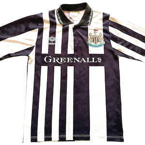 Pode incluir: Uma camisa de futebol listrada em preto e branco com gola azul e um logotipo de equipe no peito. A camisa tem o texto "GREENALLS" impresso na frente. O logotipo &eacute; um bras&atilde;o preto e branco com o texto "NEWCASTLE UNITED" dentro.