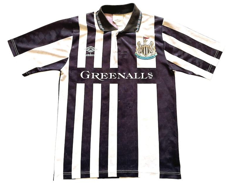 Pode incluir: Uma camisa de futebol listrada em preto e branco com gola azul e um logotipo de equipe no peito. A camisa tem o texto "GREENALLS" impresso na frente. O logotipo &eacute; um bras&atilde;o preto e branco com o texto "NEWCASTLE UNITED" dentro.
