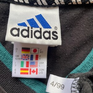 May include: Black and teal striped shirt with a white adidas logo tag and size tag. The size tag shows the size in different countries, including 42/44 in the UK, 7 in the US, and L in Canada. The shirt also has a white tag with the number 4/99.
