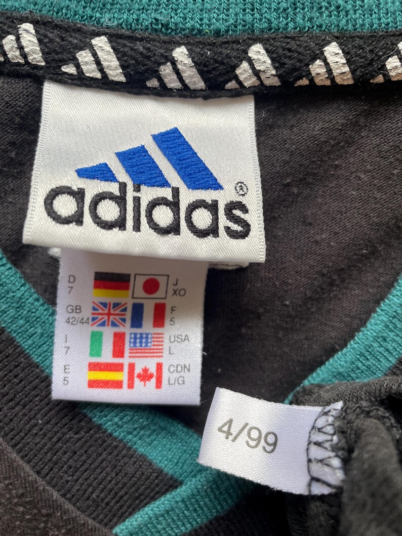 May include: Black and teal striped shirt with a white adidas logo tag and size tag. The size tag shows the size in different countries, including 42/44 in the UK, 7 in the US, and L in Canada. The shirt also has a white tag with the number 4/99.