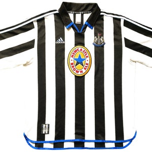 Może przedstawiać: Biało-czarna w pasy koszulka piłkarska z niebieskimi lam&oacute;wkami. Koszulka ma logo Adidasa na lewej piersi i logo Newcastle Brown Ale na prawej piersi.