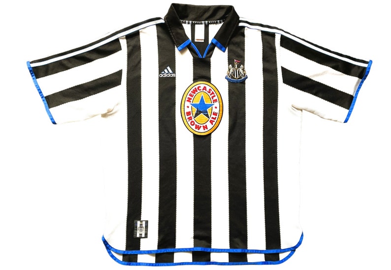 Może przedstawiać: Biało-czarna w pasy koszulka piłkarska z niebieskimi lam&oacute;wkami. Koszulka ma logo Adidasa na lewej piersi i logo Newcastle Brown Ale na prawej piersi.