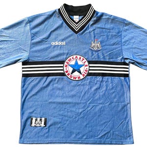 May include: A light blue soccer jersey with black and white stripes across the chest. The jersey has the Adidas logo on the left chest and the Newcastle United crest on the right chest. The crest is surrounded by a circle with the words "Newcastle Brown Ale" in red and white.