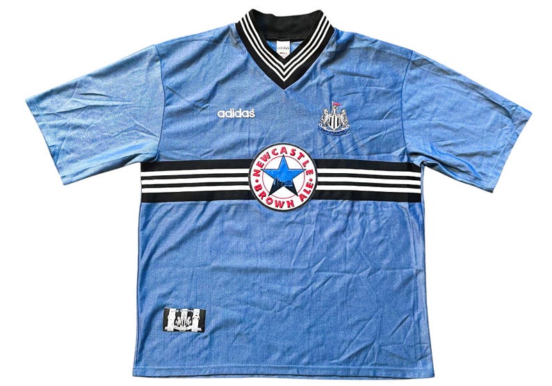 May include: A light blue soccer jersey with black and white stripes across the chest. The jersey has the Adidas logo on the left chest and the Newcastle United crest on the right chest. The crest is surrounded by a circle with the words "Newcastle Brown Ale" in red and white.