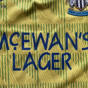 May include: A yellow and green soccer jersey with a blue and white logo that says "Newcastle United". The jersey has a blue sponsor logo that says "McEwans' Lager".