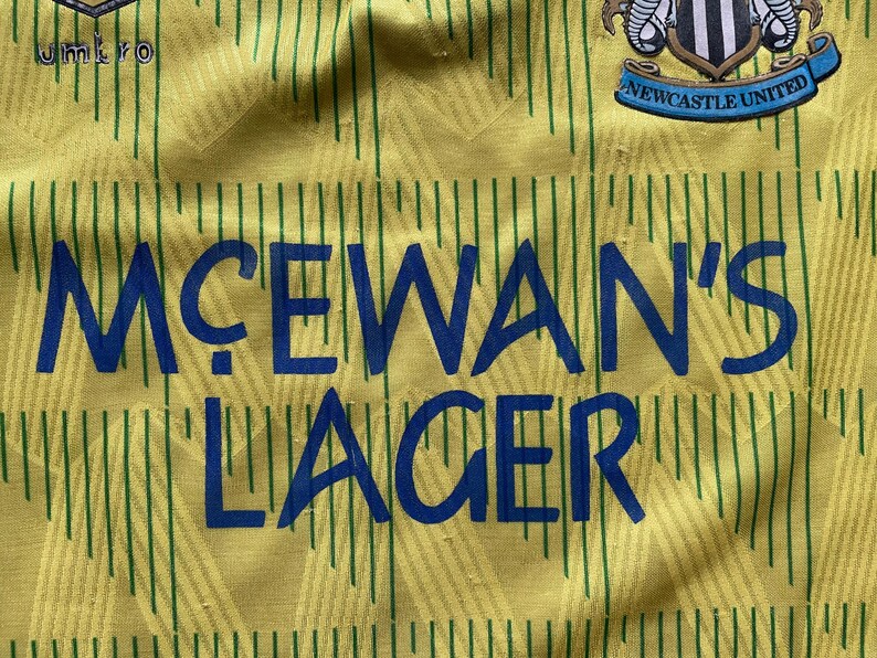 May include: A yellow and green soccer jersey with a blue and white logo that says "Newcastle United". The jersey has a blue sponsor logo that says "McEwans' Lager".