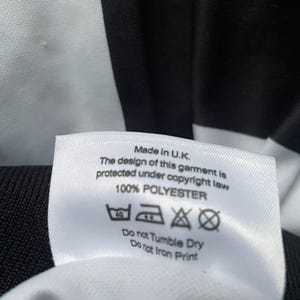 May include: Close-up of a white label with black text, indicating the garment is made in the U.K. and is 100% polyester. The label also includes care instructions: wash at 40 degrees, do not tumble dry, and do not iron print.