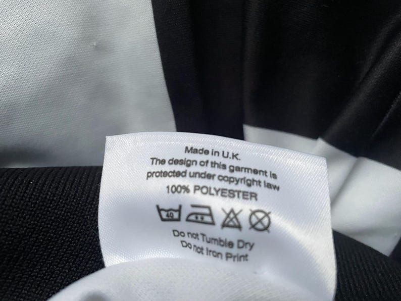May include: Close-up of a white label with black text, indicating the garment is made in the U.K. and is 100% polyester. The label also includes care instructions: wash at 40 degrees, do not tumble dry, and do not iron print.