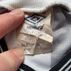 May include: White and black striped soccer jersey with a white tag that says "Umbro" and "86-91cm 34/38inch MADE IN ENGLAND".