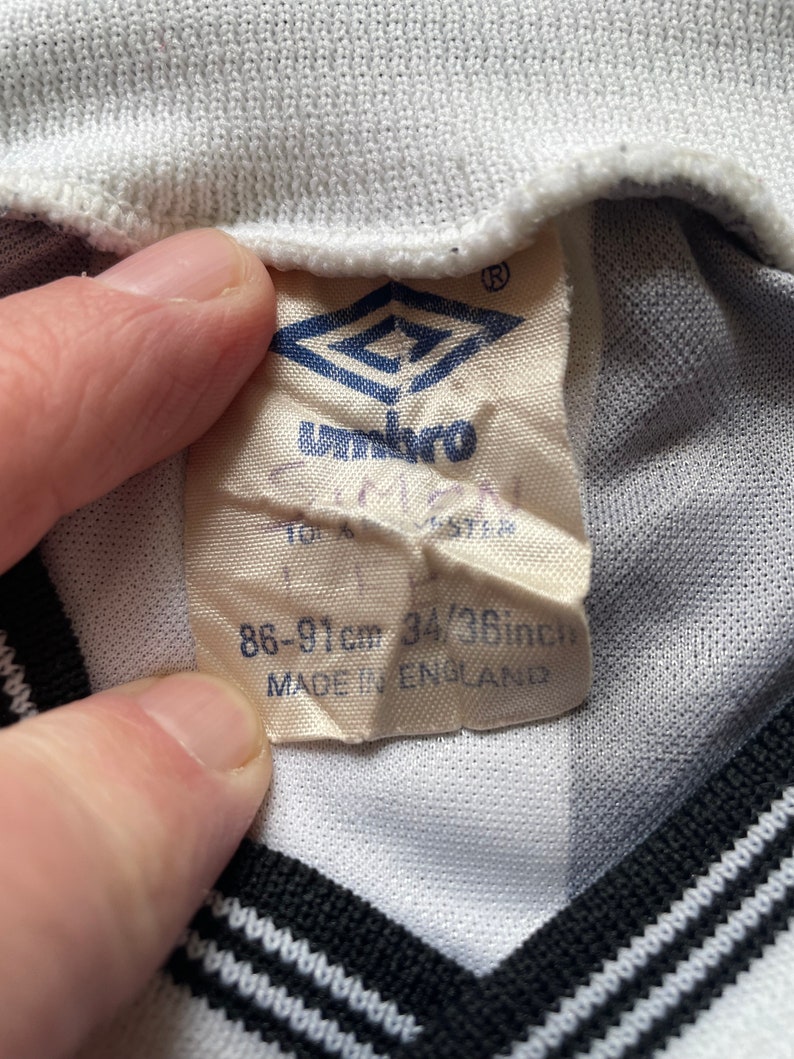 May include: White and black striped soccer jersey with a white tag that says "Umbro" and "86-91cm 34/38inch MADE IN ENGLAND".