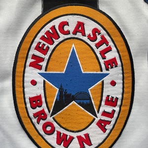 May include: Close-up of a white and black striped jersey with a Newcastle Brown Ale logo. The logo is a circular emblem with the words "Newcastle Brown Ale" in red, surrounding a blue star with a city silhouette.