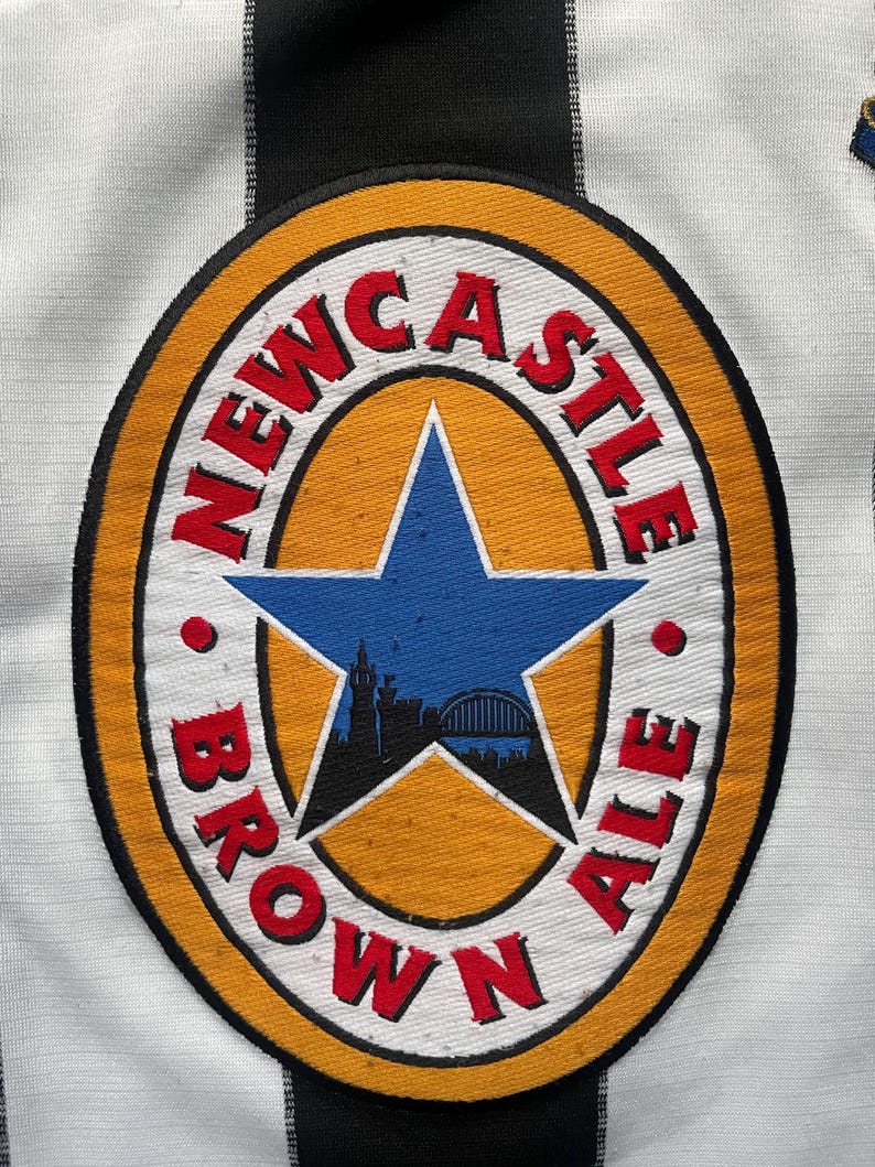May include: Close-up of a white and black striped jersey with a Newcastle Brown Ale logo. The logo is a circular emblem with the words "Newcastle Brown Ale" in red, surrounding a blue star with a city silhouette.