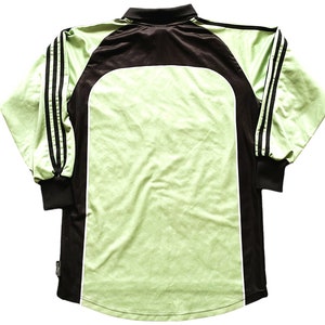 May include: A long-sleeved athletic jersey in shades of green and black. The jersey features a light green body with black accents on the shoulders, sleeves, and collar. Three black stripes run down each sleeve. The jersey has a classic athletic design.