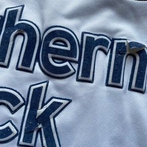 May include: Close-up of a white jersey with the text "Southern" and "ock" in navy blue lettering.