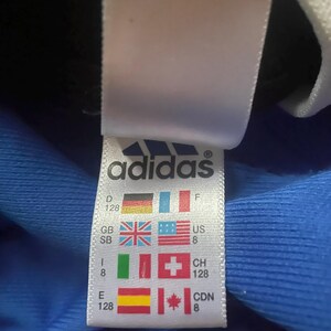 K&ouml;nnte beinhalten: Ein wei&szlig;es Adidas-Etikett mit dem Markenlogo und L&auml;nderflaggen, einschlie&szlig;lich der deutschen Flagge. Das Etikett zeigt auch Gr&ouml;&szlig;eninformationen.