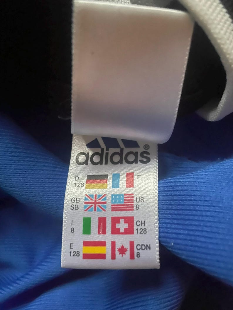 K&ouml;nnte beinhalten: Ein wei&szlig;es Adidas-Etikett mit dem Markenlogo und L&auml;nderflaggen, einschlie&szlig;lich der deutschen Flagge. Das Etikett zeigt auch Gr&ouml;&szlig;eninformationen.
