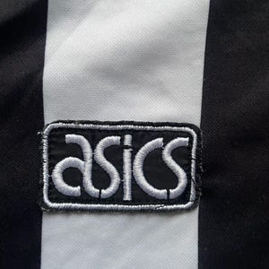May include: Close-up of a black and white striped fabric with an embroidered ASICS logo patch. The logo is white on a black background, with a white border. The fabric has vertical black and white stripes.